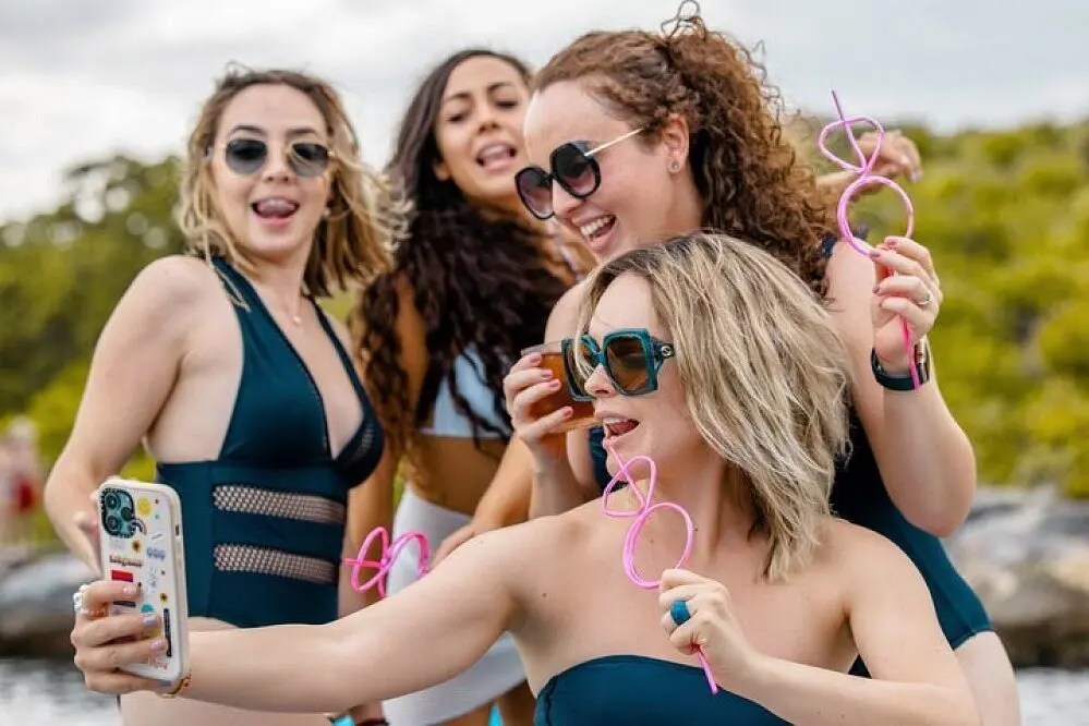 Four women in swimsuits and sunglasses taking a selfie with novelty pink drinking straws