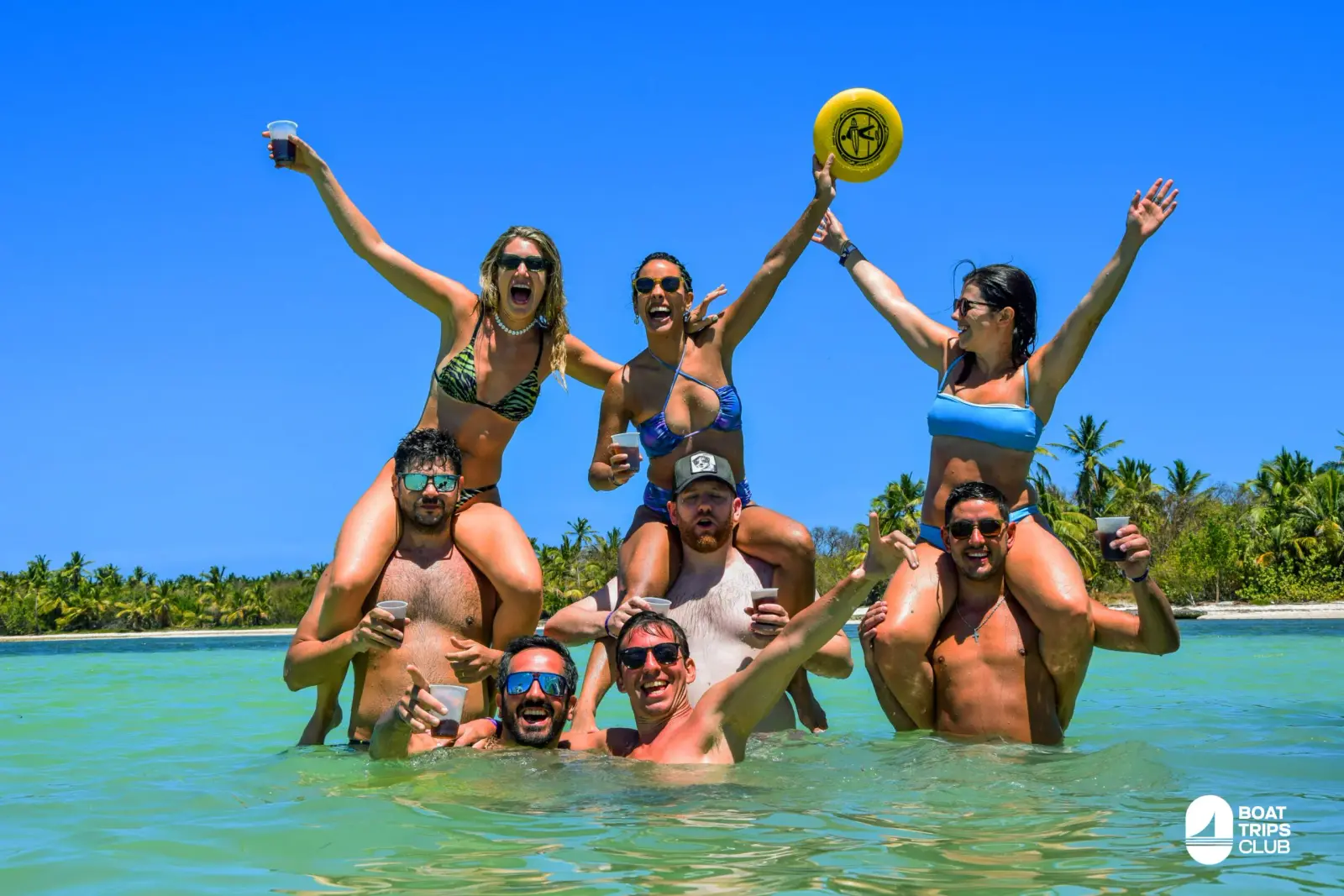 Three women ride on friends' shoulders in the shallows off a palm-lined Punta Cana beach, cheering with drinks