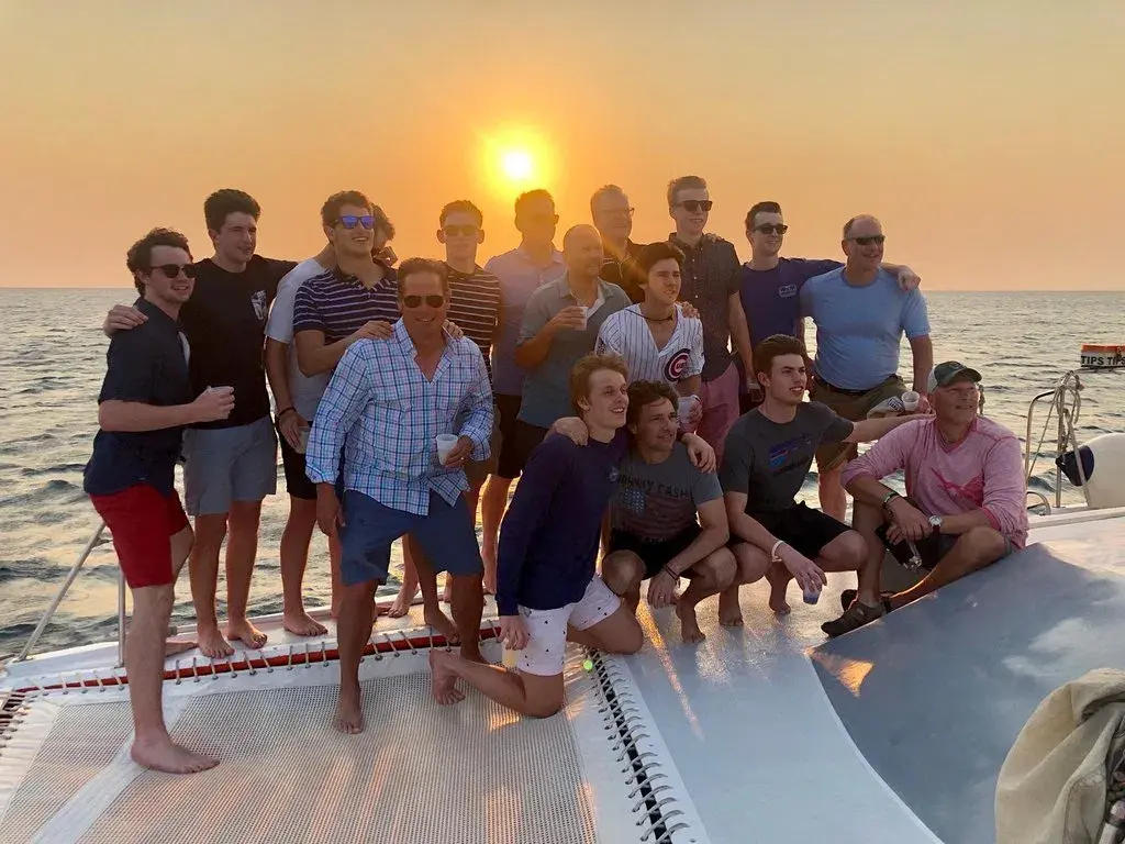 Large group of guests posing together on the catamaran's front deck as the sun sets off Aruba