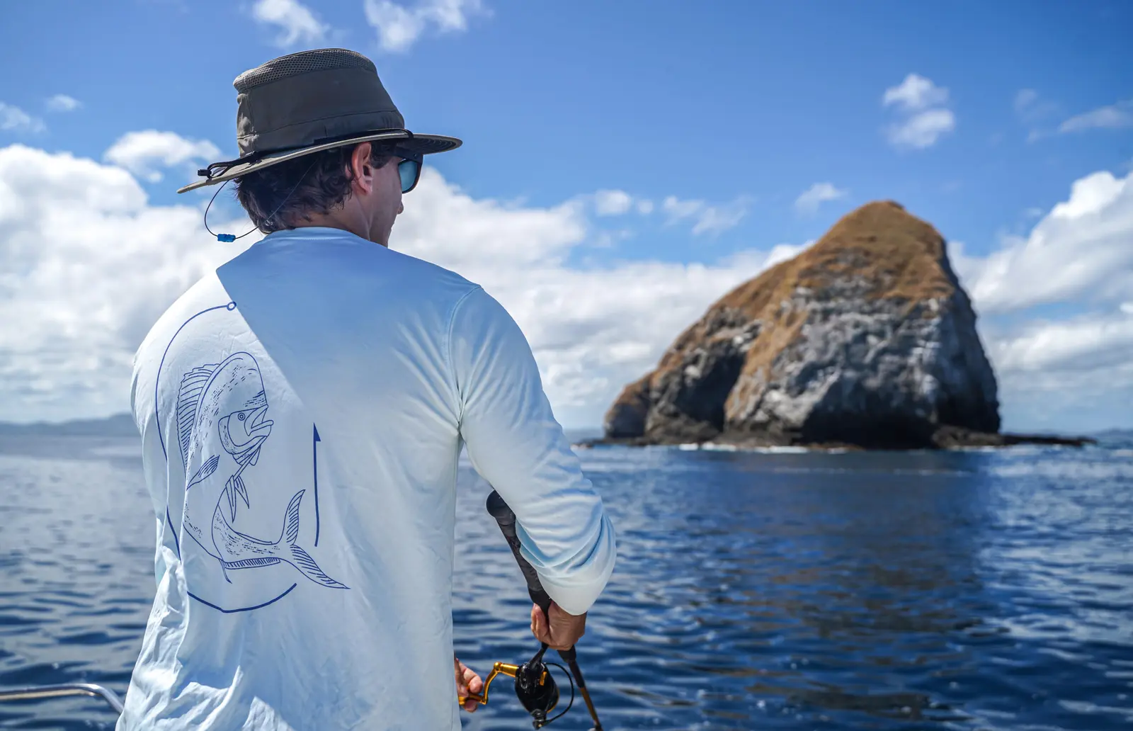 An angler in a sun hat holds his rod while facing a towering rock islet off the coast of Costa Rica