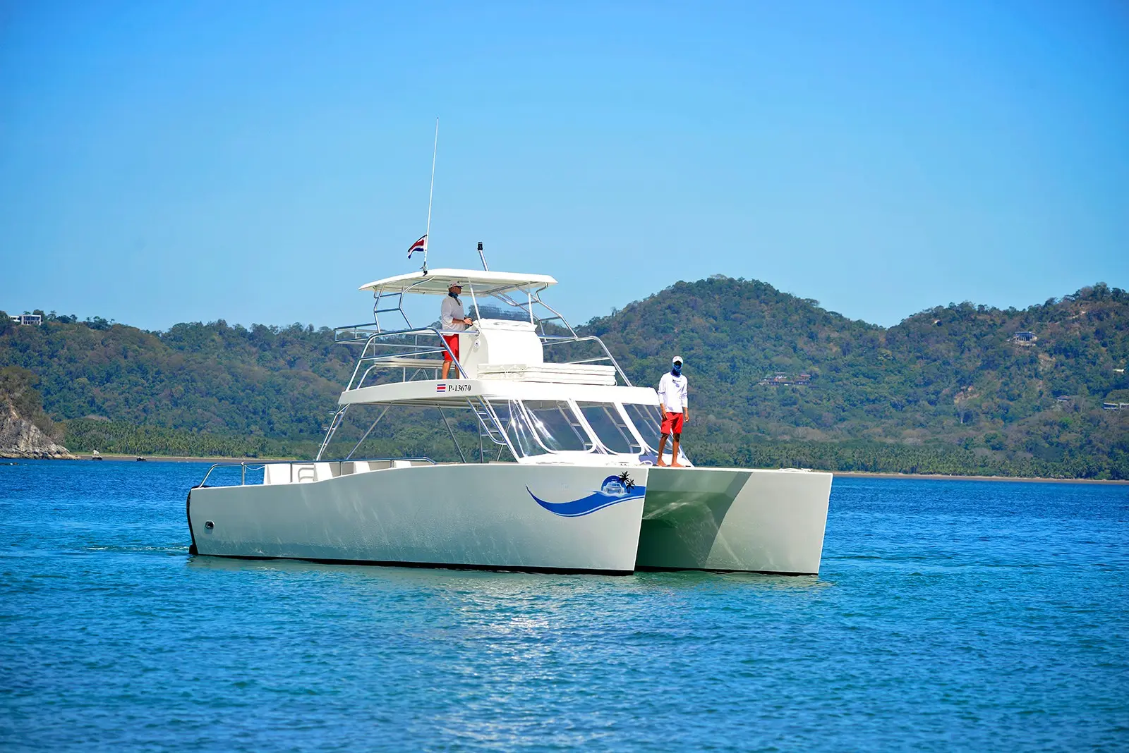 Crew standing on the bow and flybridge of a white catamaran on blue Gulf of Nicoya water below green hills