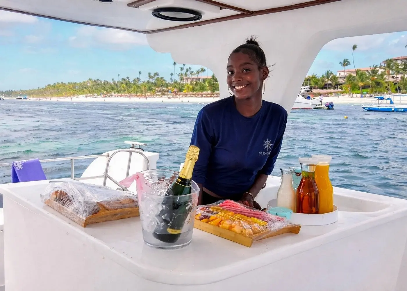 Smiling crew member at the catamaran bar with sparkling wine on ice and a snack tray, palm beach behind