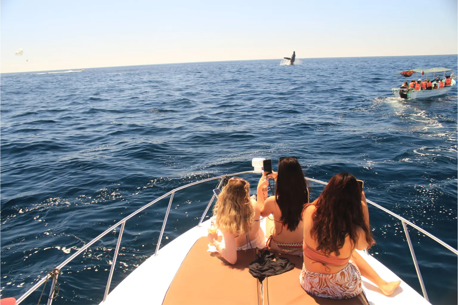 Three women on a yacht's bow watching a whale breach in the distance off Cabo San Lucas