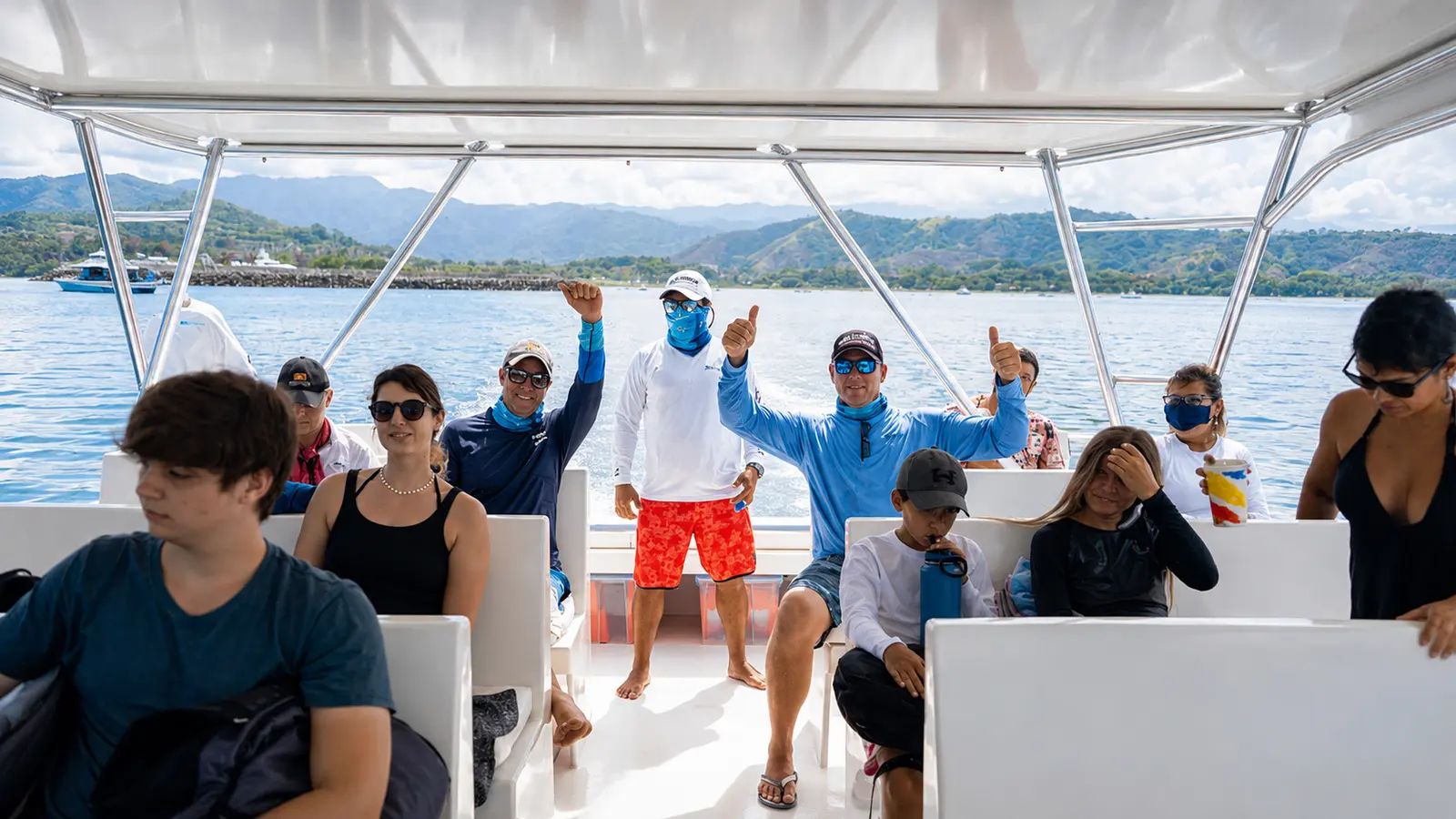 Guests, some in face masks, waving from the shaded deck of a catamaran with Costa Rica's green hills behind
