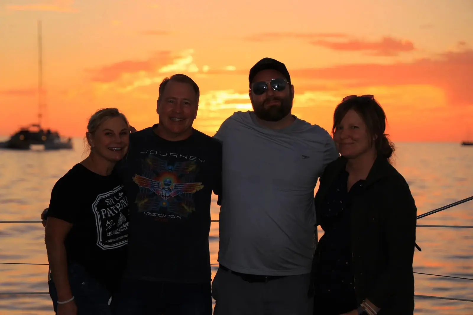 Four guests pose on the catamaran deck against an orange sunset sky with a sailboat behind