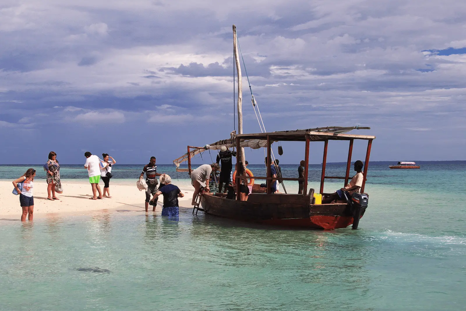 Guests stepping off a wooden dhow onto a white sandbank under a cloudy sky off Zanzibar
