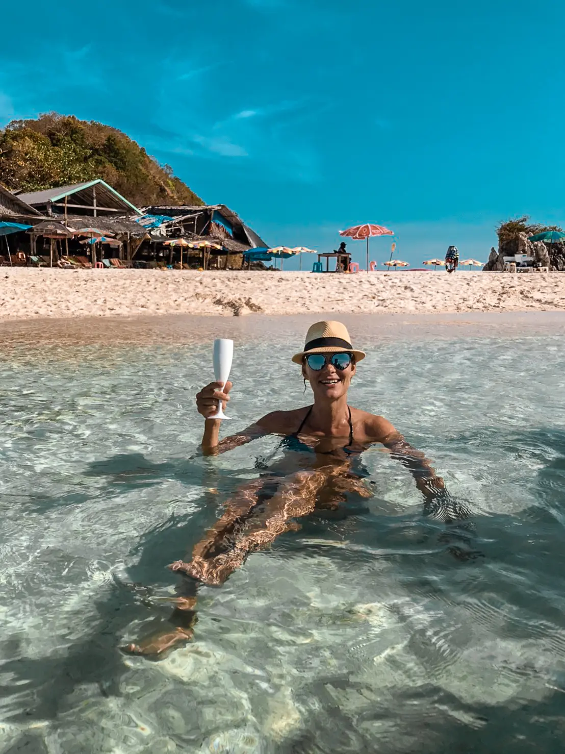 Woman in a sun hat sitting in clear shallows and raising a bottle, beach huts on a Khai island behind