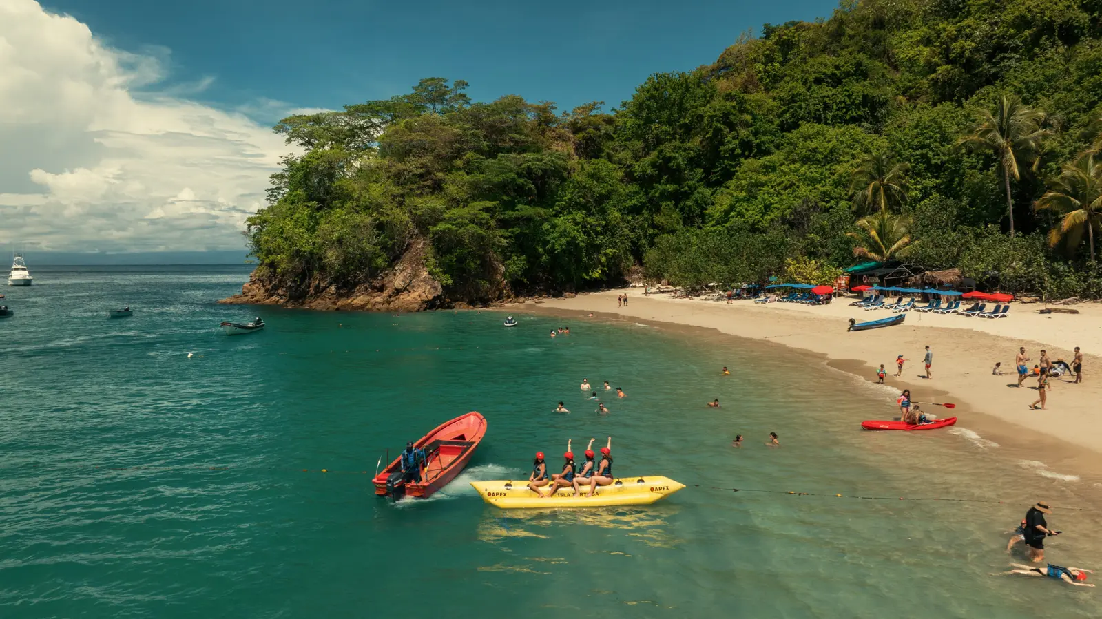 Yellow banana boat full of riders pulled by a red inflatable past swimmers at a Tortuga Island beach
