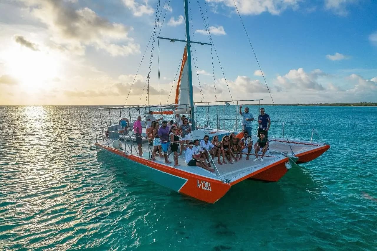 Guests crowding the deck of an orange-hulled sailing catamaran on turquoise water off Aruba