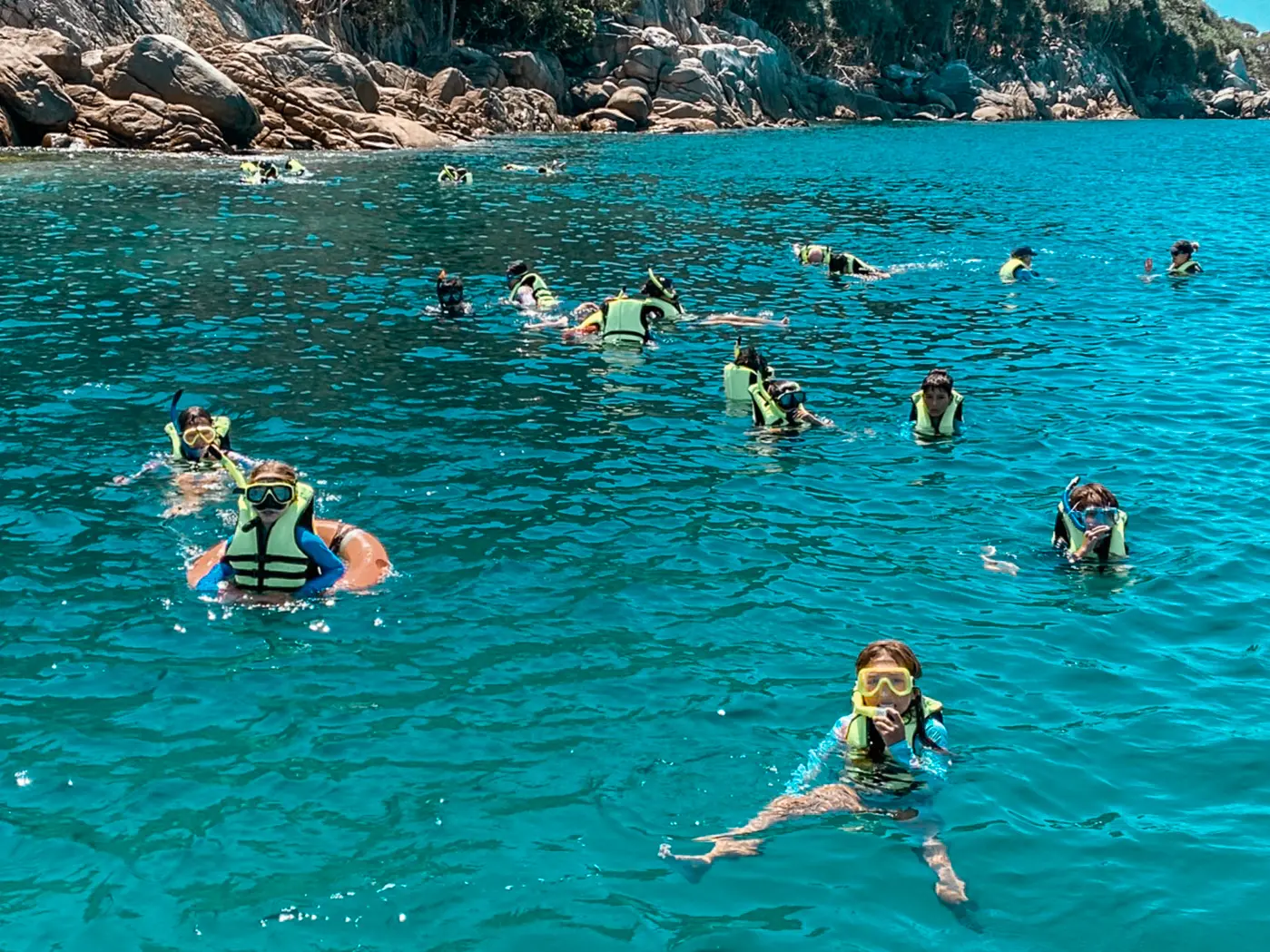 Large group of snorkelers in life vests and masks spread across turquoise water off a rocky Racha shore