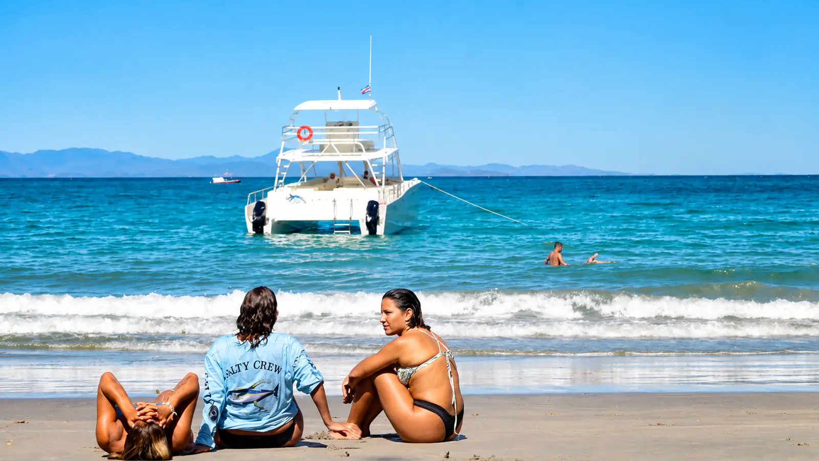 Three guests relaxing on the sand at Tortuga Island with the catamaran anchored past the surf