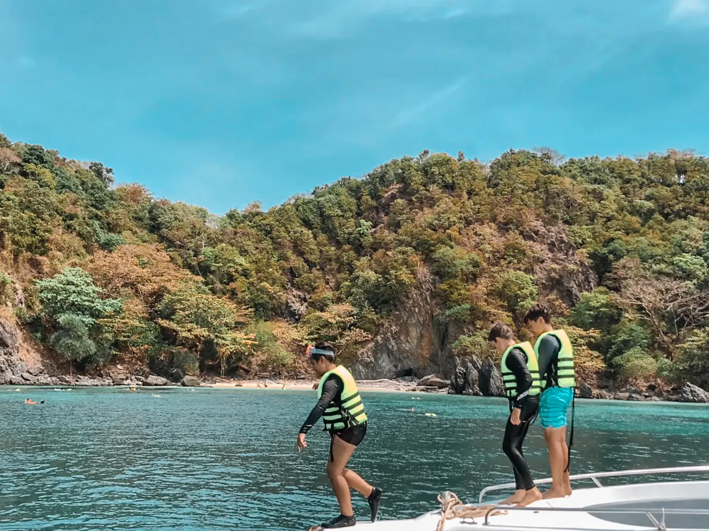 Three guests in life vests standing on the speedboat bow off a jungle-covered island in Thailand