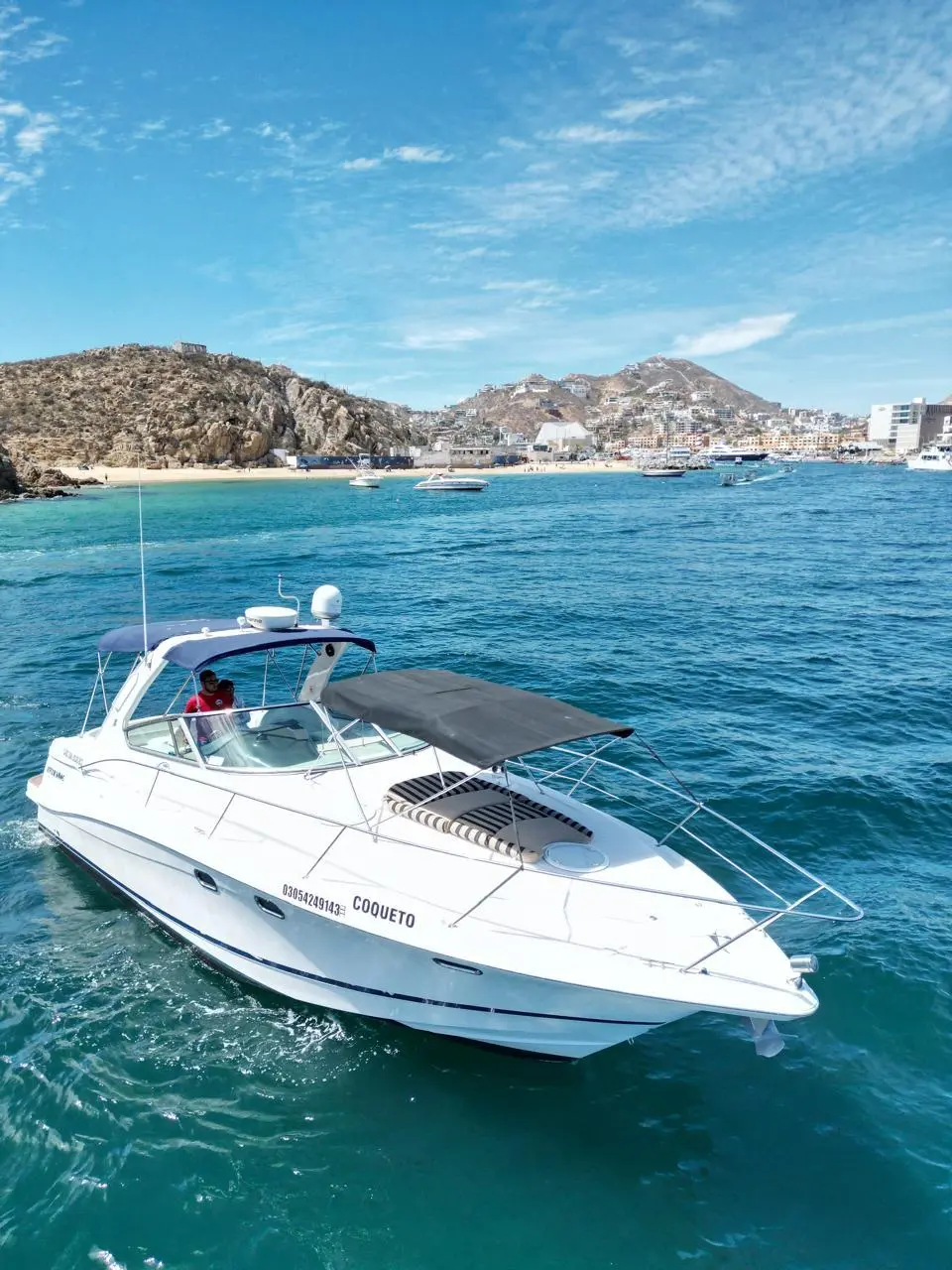 Yacht anchored in clear teal water below rocky hills and hotels of Cabo San Lucas