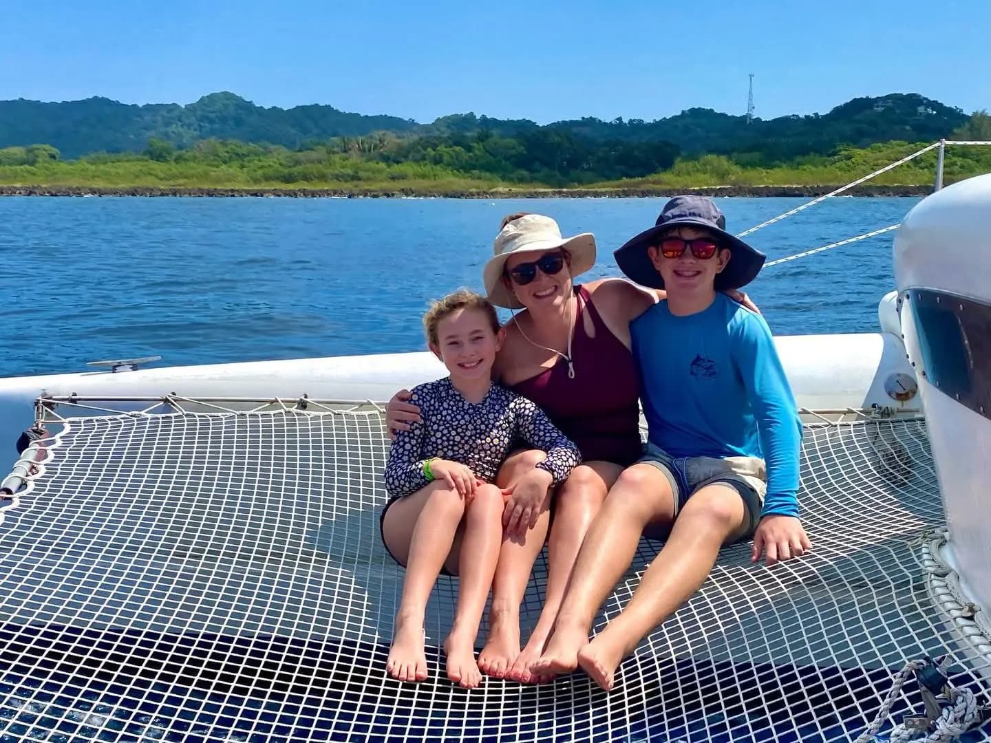 A woman and two kids sitting on the catamaran's trampoline net with green Roatan hills behind
