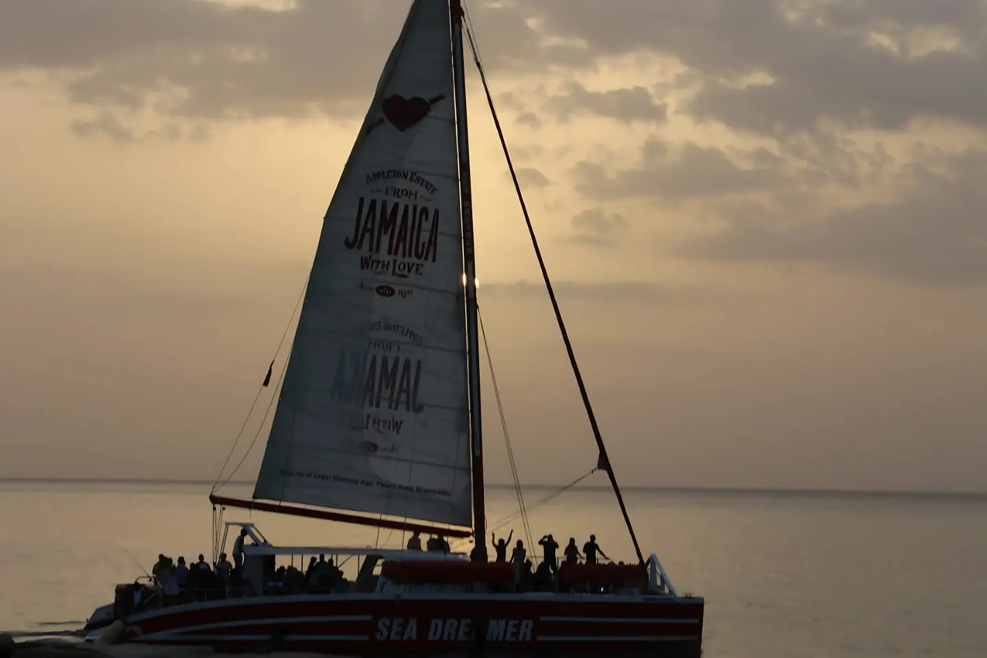 Silhouette of a sailing catamaran with guests on deck and a raised mainsail at dusk off Jamaica