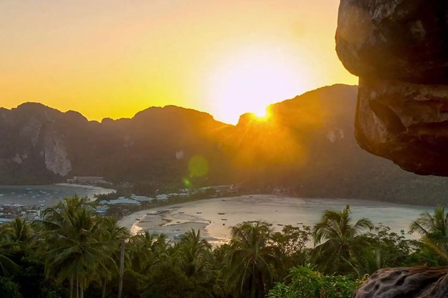 Sun sinking behind jungle-covered peaks above a curved bay and palm trees, seen from a Thailand viewpoint