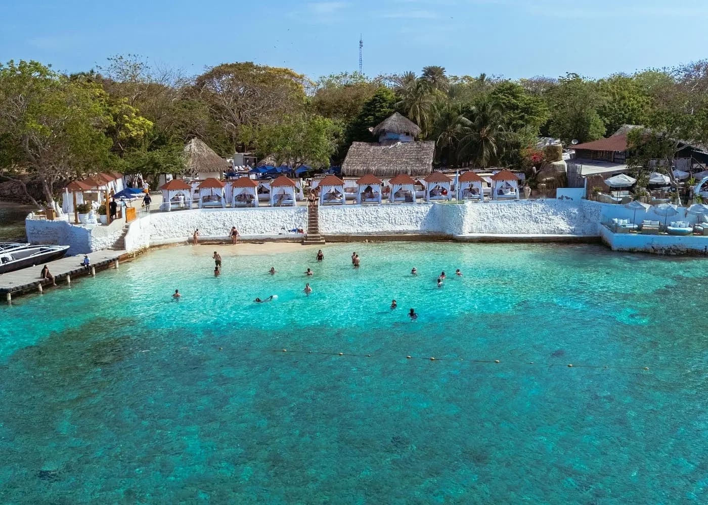 Aerial view of an island beach club with white cabanas above a turquoise cove full of swimmers near Cartagena