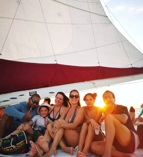 Smiling guests and a child posing under a catamaran's sail with the low sun glowing behind them