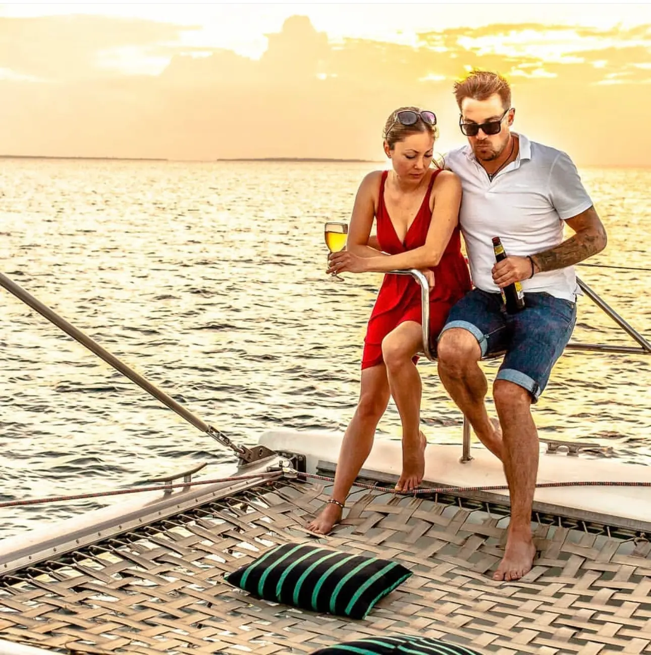 Couple with drinks sitting on the bow net of a catamaran in golden sunset light off Cancún