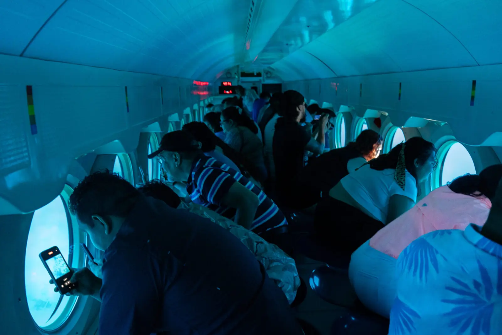 Passengers peering through round portholes inside a submarine cabin, one taking a phone photo