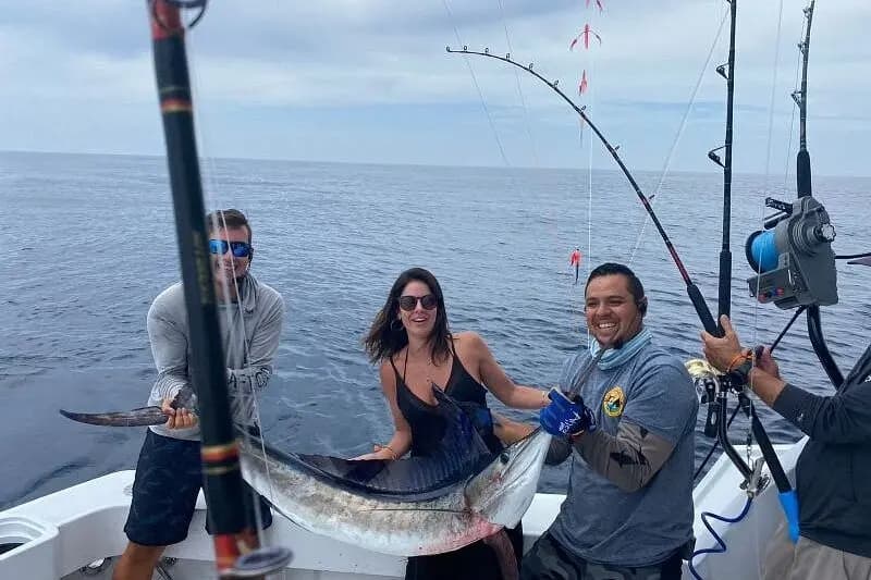 A woman and two men hold up a striped marlin among the rods in the boat's cockpit