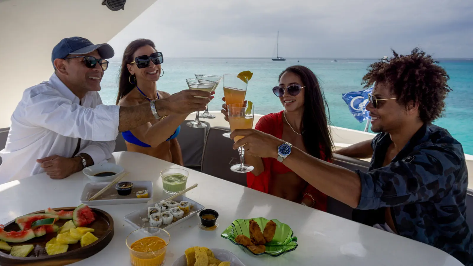 Four guests toasting cocktails over a table of fruit, sushi and snacks on the yacht's deck