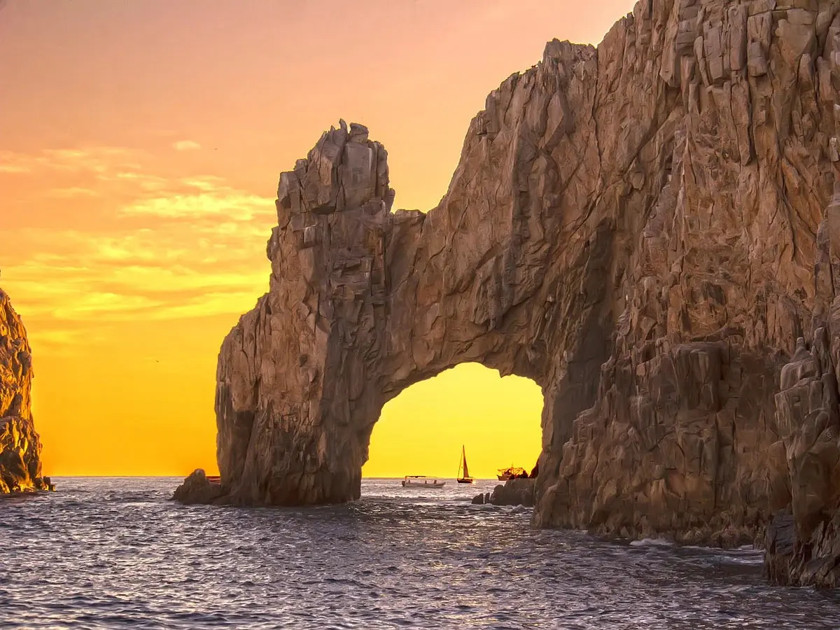 El Arco silhouetted against a golden sunset sky with a sailboat visible through the arch