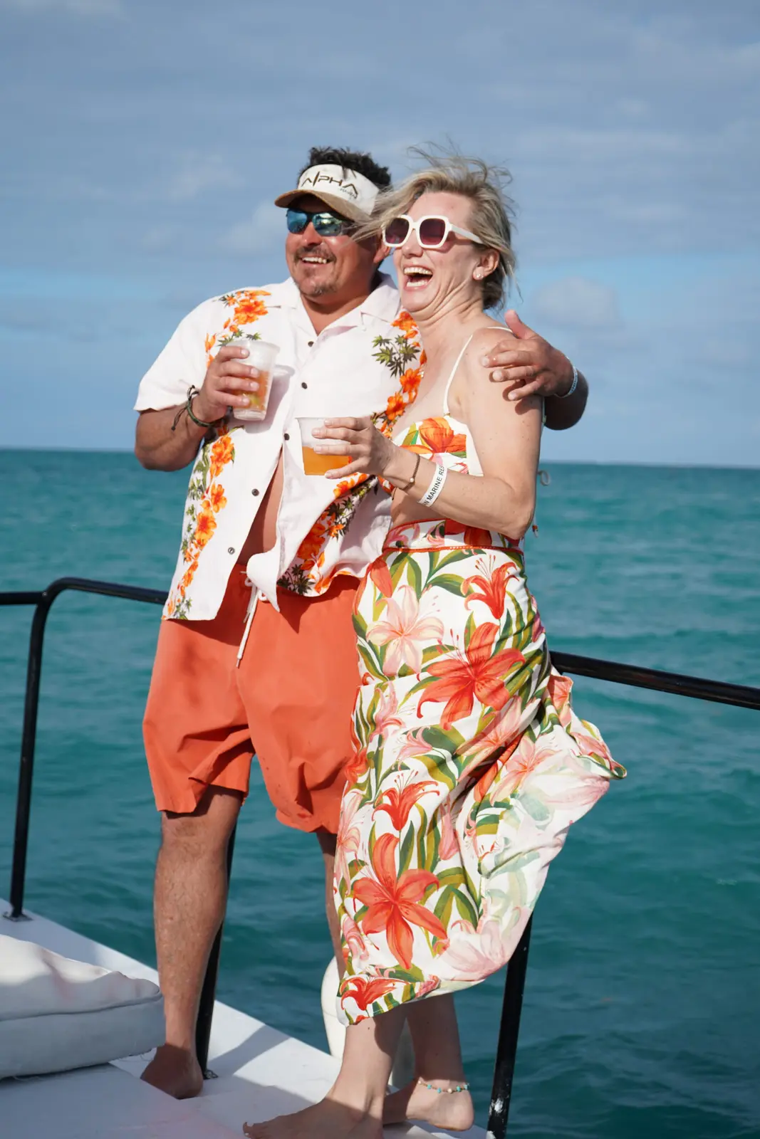 Couple in matching tropical-print outfits laughing with drinks on a catamaran deck