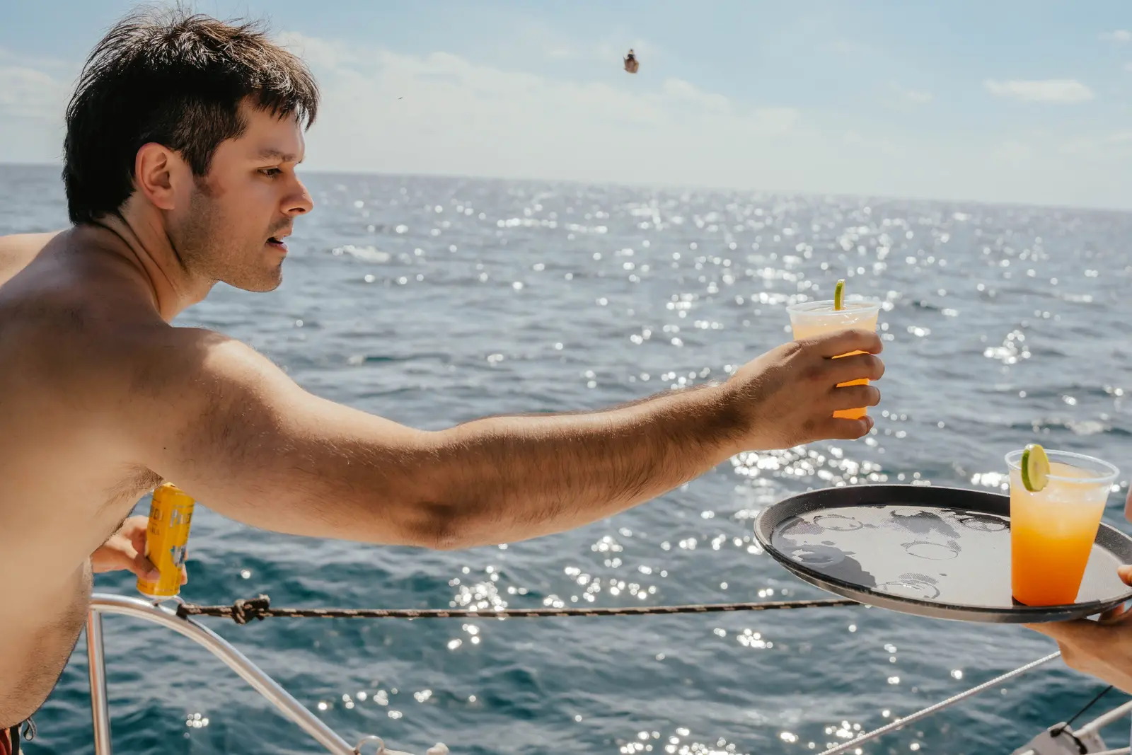 Shirtless guest taking a drink from a tray of cocktails at a yacht's rail above sparkling sea