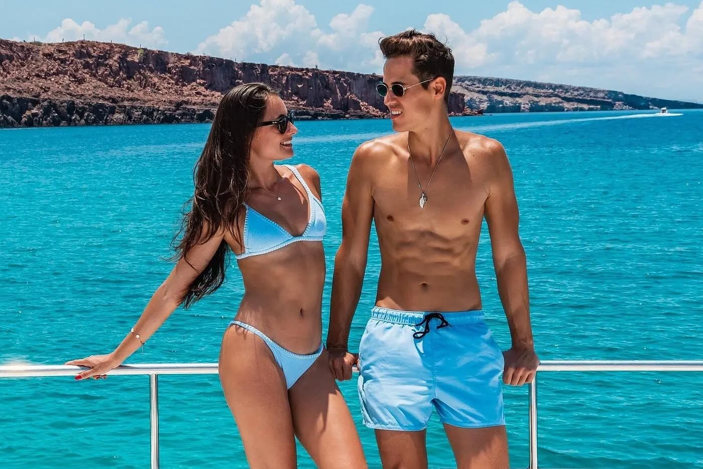 Couple in matching blue swimwear at the boat rail with red rocky cliffs behind
