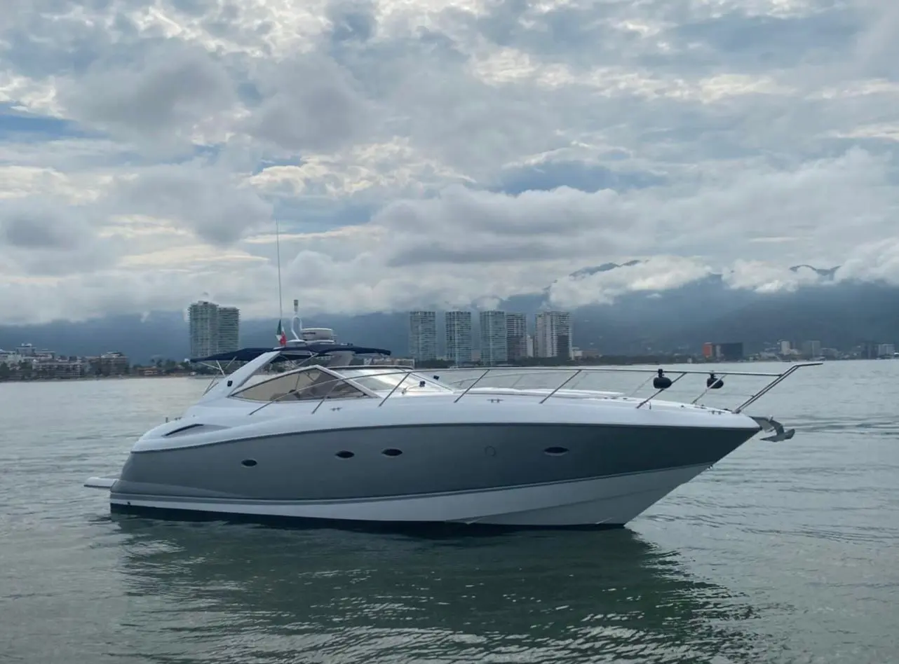 Sleek white motor yacht on calm water with high-rise towers and cloud-covered hills behind