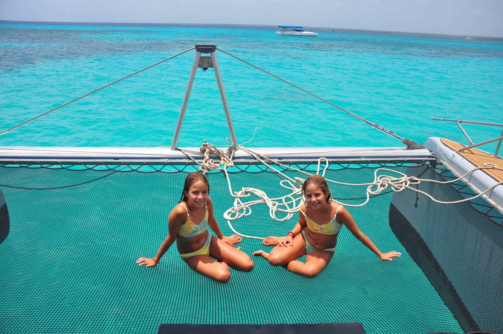 Two girls in matching swimsuits sit on the trampoline net at the bow of the power catamaran