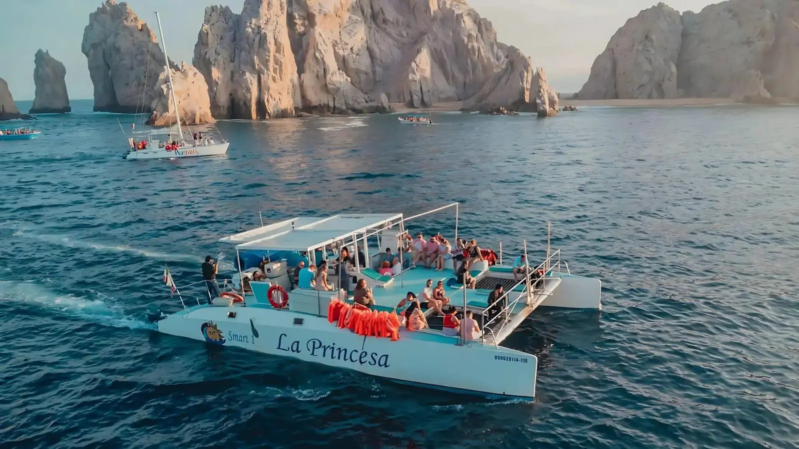 Catamaran full of guests cruising past the rock formations of Cabo San Lucas, a sailboat in the distance