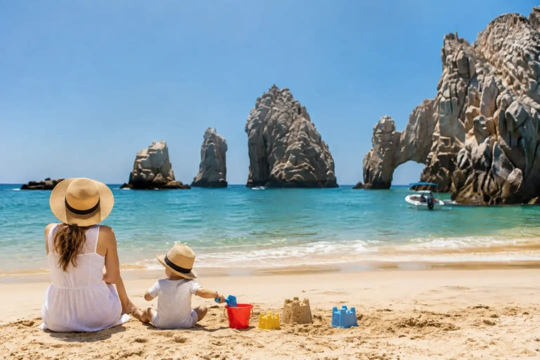 Woman beside a child building sandcastles on a Cabo San Lucas beach, facing the rock arch and a motorboat