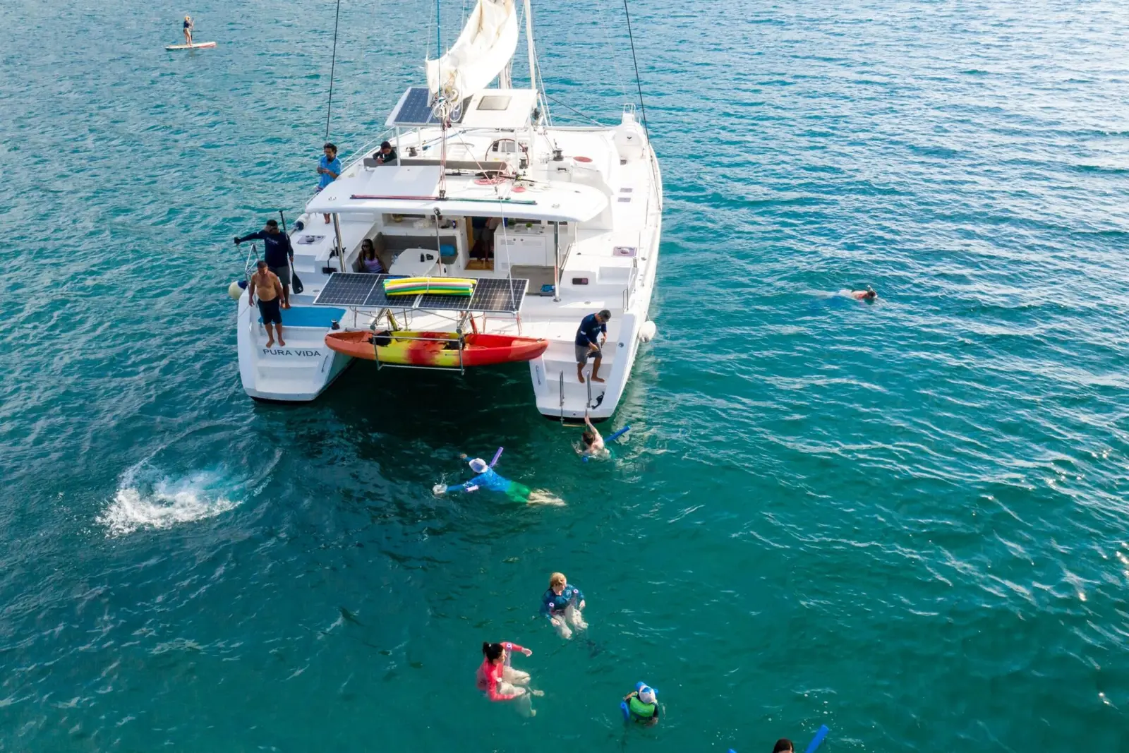 Guests swimming and snorkeling around the stern of an anchored sailing catamaran with a kayak aboard