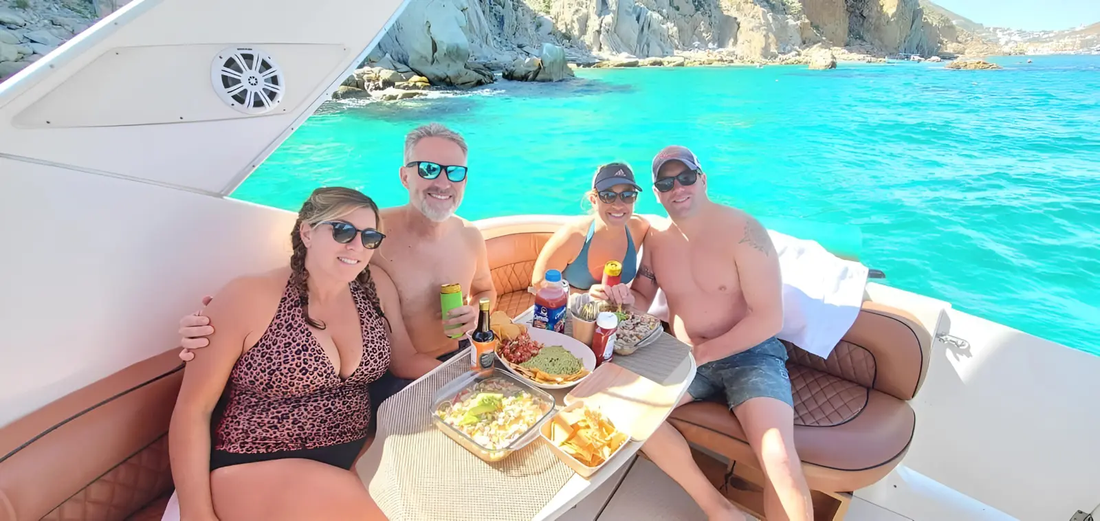 Two couples sharing snacks and drinks at the cockpit table of a yacht in a turquoise Cabo San Lucas cove