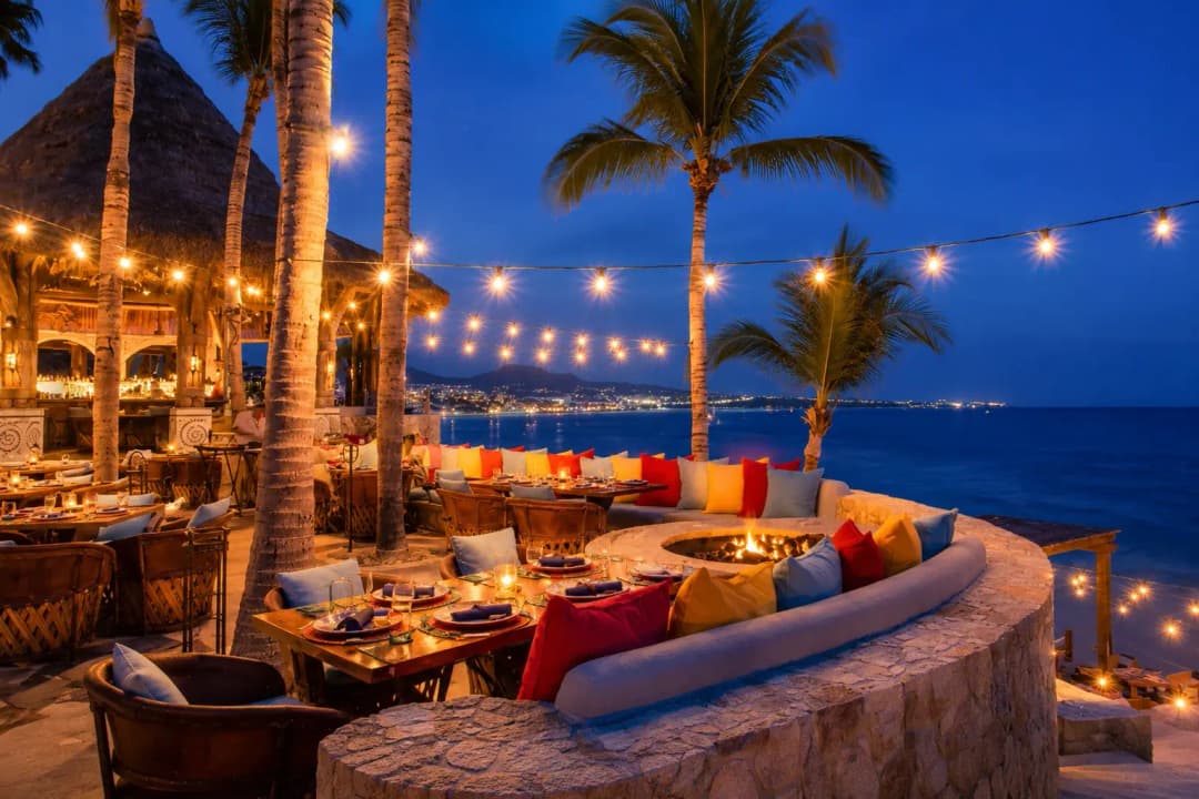 Oceanfront restaurant in Cabo San Lucas with palm trees, colorful seating, fire pit, and warm string lights at twilight.