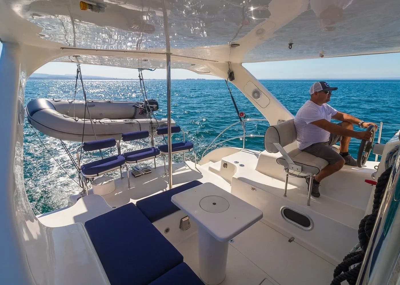 A captain steering from the helm seat of a catamaran, a dinghy hanging off the stern