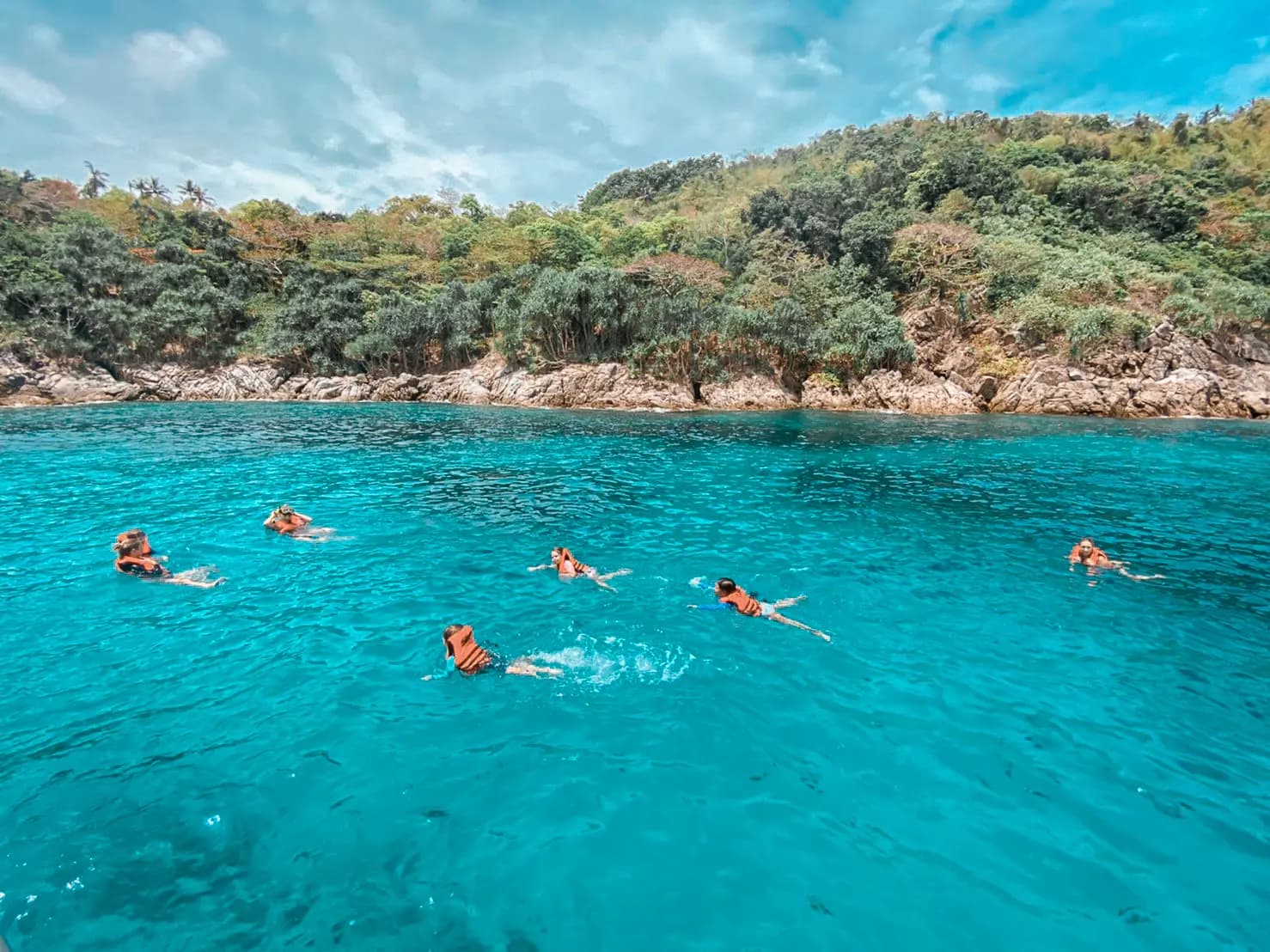 Guests snorkeling in clear turquoise water off a rocky, forested island shore in Thailand