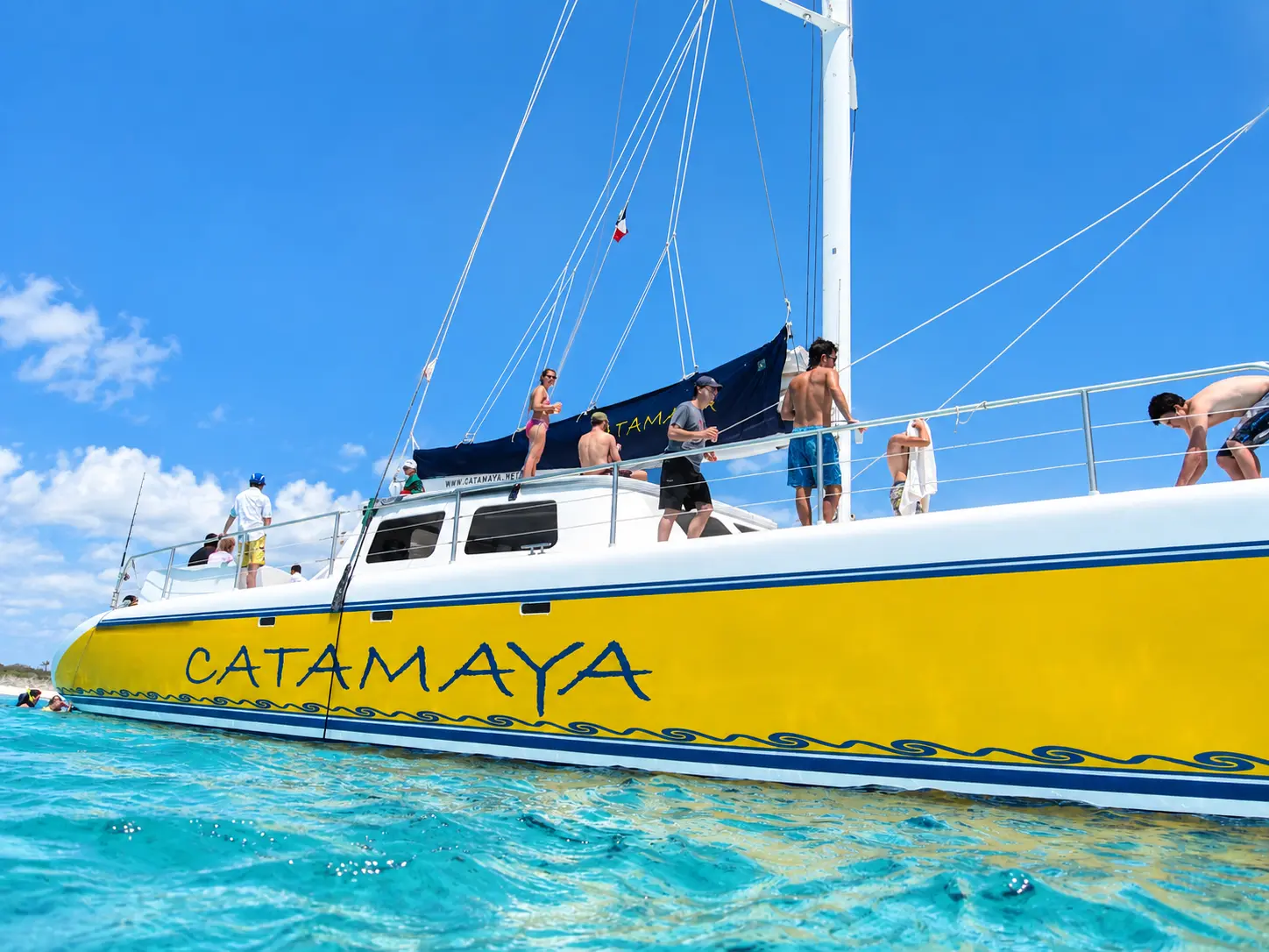 Guests along the rail of a yellow sailing catamaran anchored in turquoise water, seen from the waterline