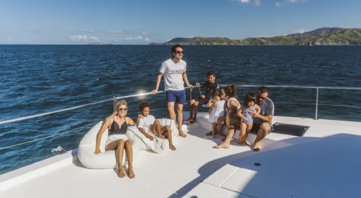 A family with kids gathers on the bow of the power catamaran, coastal hills across the water