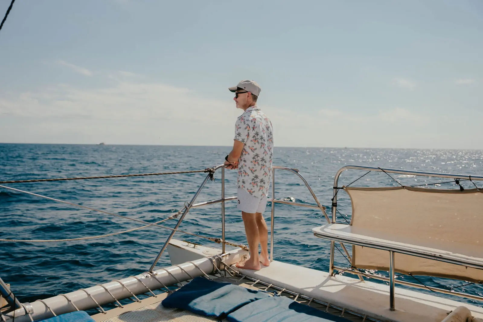 Man in a floral shirt and cap at a catamaran's bow rail gazing out over the sea off Los Cabos