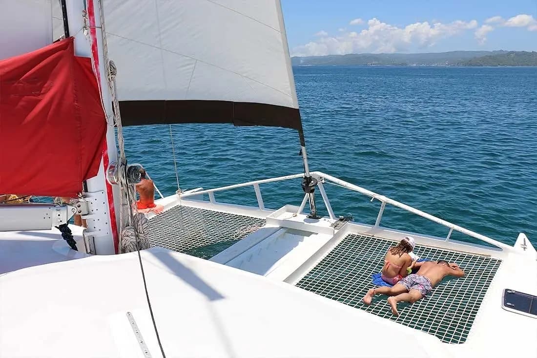 Couple lying on the trampoline net of a sailing catamaran beneath red and white sails off Samaná