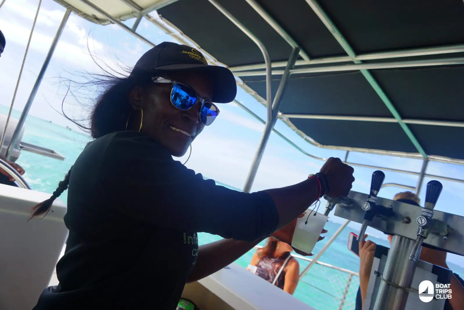 Crew member in a cap and mirrored sunglasses pouring a drink from the taps at the catamaran's onboard bar