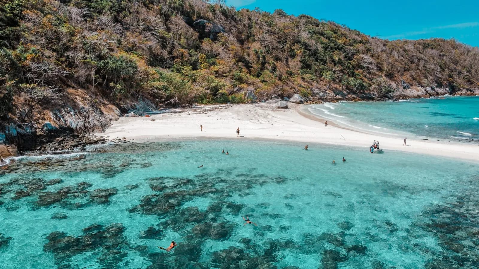 White sand bay with dark reef patches in turquoise water below a wooded hillside on Racha Island