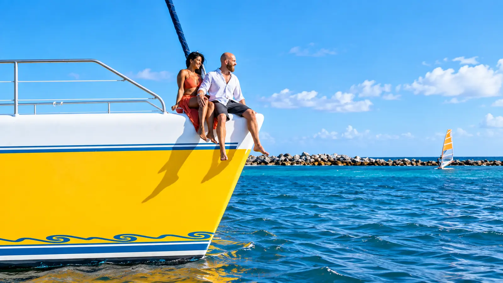 Couple sitting on the bow of the yellow catamaran, a rock breakwater and small sailboat beyond