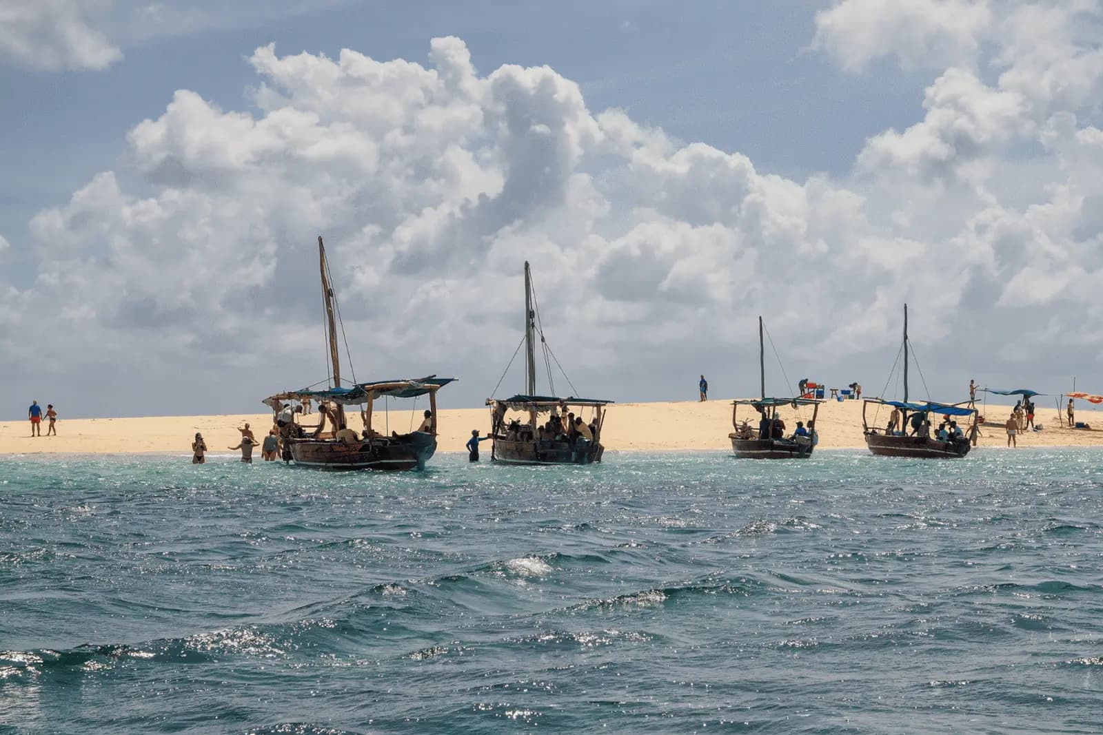 Four wooden dhows with tall masts anchored off a Zanzibar sandbank under towering white clouds
