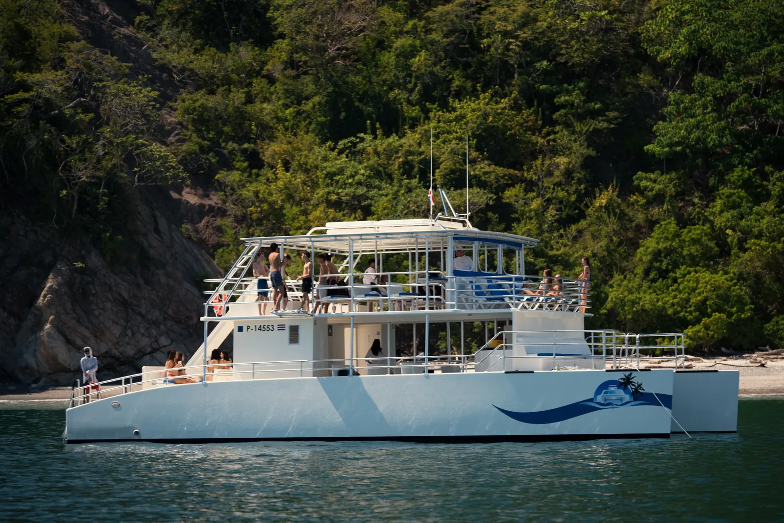 Two-deck catamaran anchored close to a forested rocky shore in the Gulf of Nicoya with guests aboard