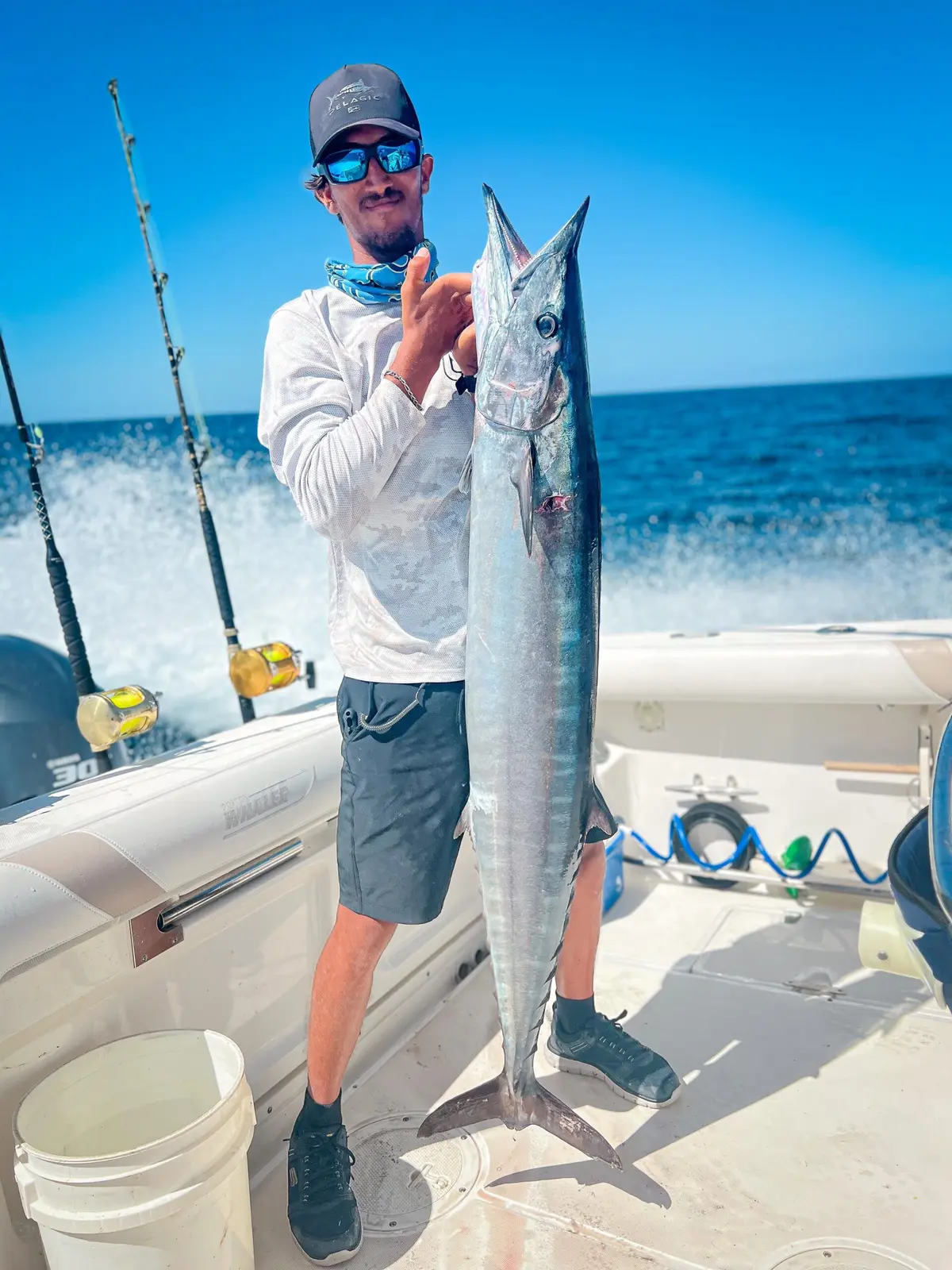 An angler holds up a long, slender silver fish on the fishing boat's deck, rods and wake behind him