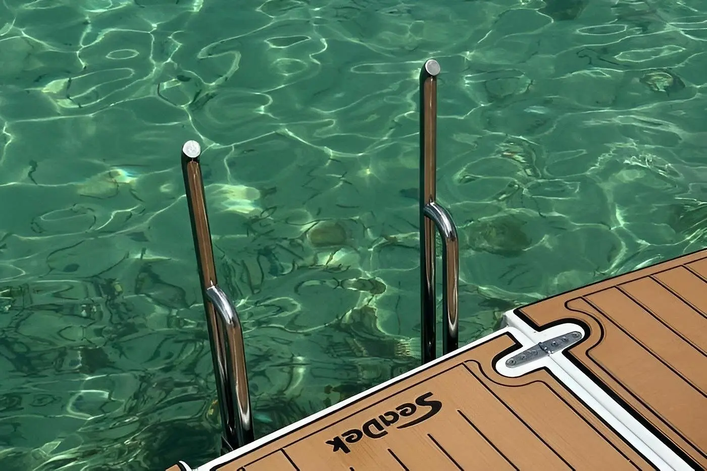 Stainless steel swim ladder dropping from a power catamaran's teak-look platform into clear green water